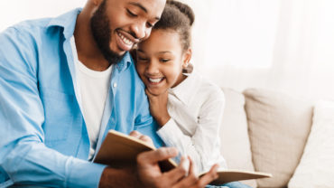 Father and daughter reading book together, sitting on sofa