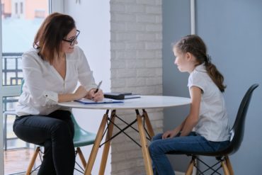 Female school psychologist and young girl sitting at round, white table talk while psychologist takes case notes.