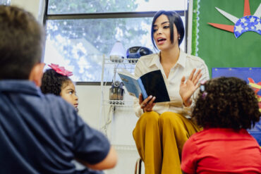Young female Hispanic schoolteacher reading aloud to elementary aged students sitting around her on the floor.