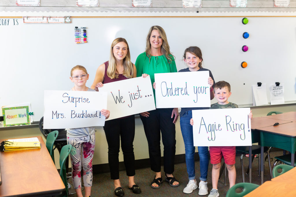 Buckland, Adams and students holding banners that read "Surprise Mrs. Buckland! We just ordered your Aggie ring!"