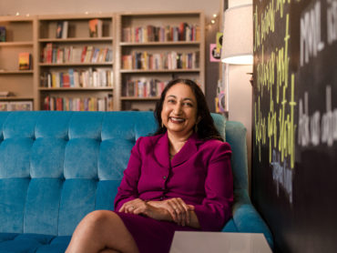 Dr. Kay Wijekumar sits on a blue velvet couch in a fuchsia suit in front of a set of book shelves
