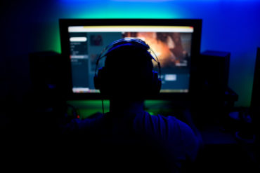 Adult man sits in front of a computer screen playing a video game, wearing headphones with green and blue LED lights creating illumination in dark room.