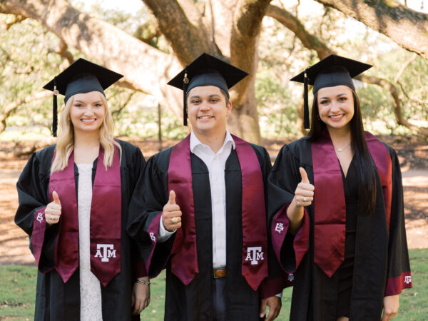 Three graduating students of Texas A&M University wearing their cap and gown while giving the "Gig 'em" thumbs up sign.