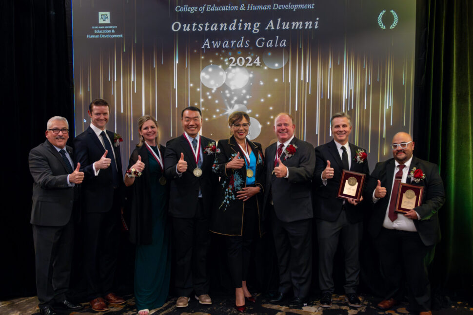 Seven honorees of Outstanding Alumni Awards Gala pose for a photo with Dean de Miranda while giving the Gig 'em thumbs up.