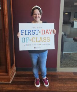 Lee holds a “First Day of Class” sign and smiles in front of a maroon wall.