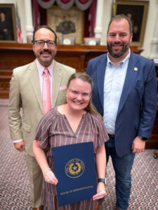Shelbi Davenport stands inside the Texas House chamber holding a blue House of Representatives folder, joined by two state officials.