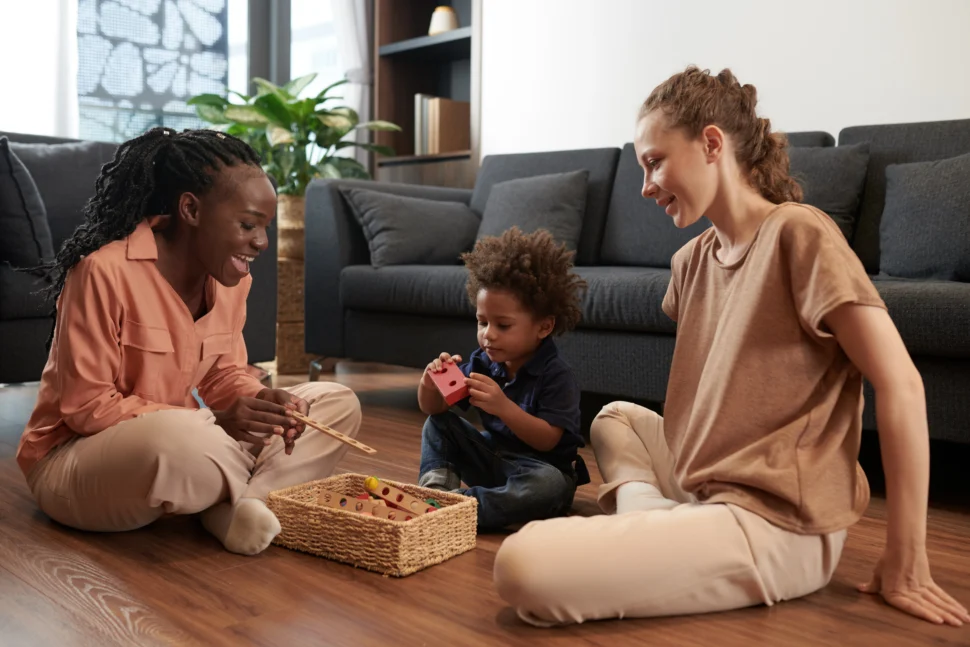 A child plays with a toy while sitting on the floor between two smiling adults. The scene is warm and cozy, with a sofa and plants in the background.