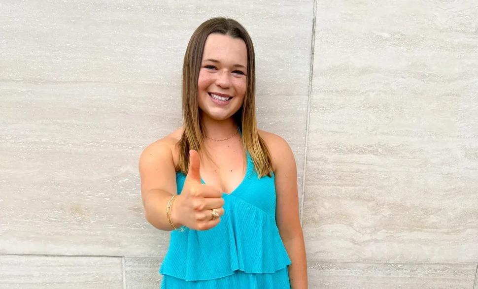 Ella Cole stands smiling against a light stone wall, wearing a teal dress and giving a Gig ’em hand sign toward the camera.