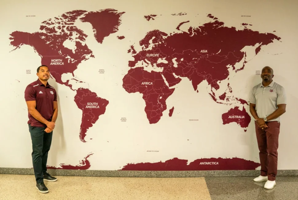 Two men stand in front of a large mural map on a wall, showing continents labeled in maroon. They are smiling, exuding a welcoming tone.