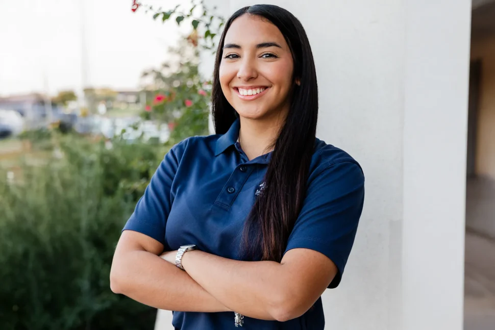 Smiling woman with long dark hair stands with arms crossed, wearing a blue polo shirt. She is outside with greenery in the background, exuding confidence.
