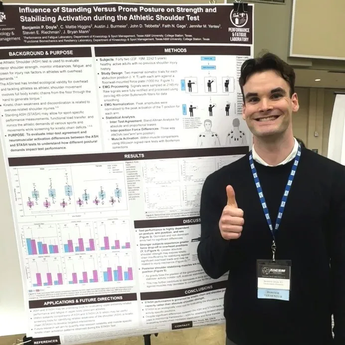 A man stands smiling and giving a thumbs-up in front of a scientific poster presentation. The poster includes graphs and text about shoulder performance.